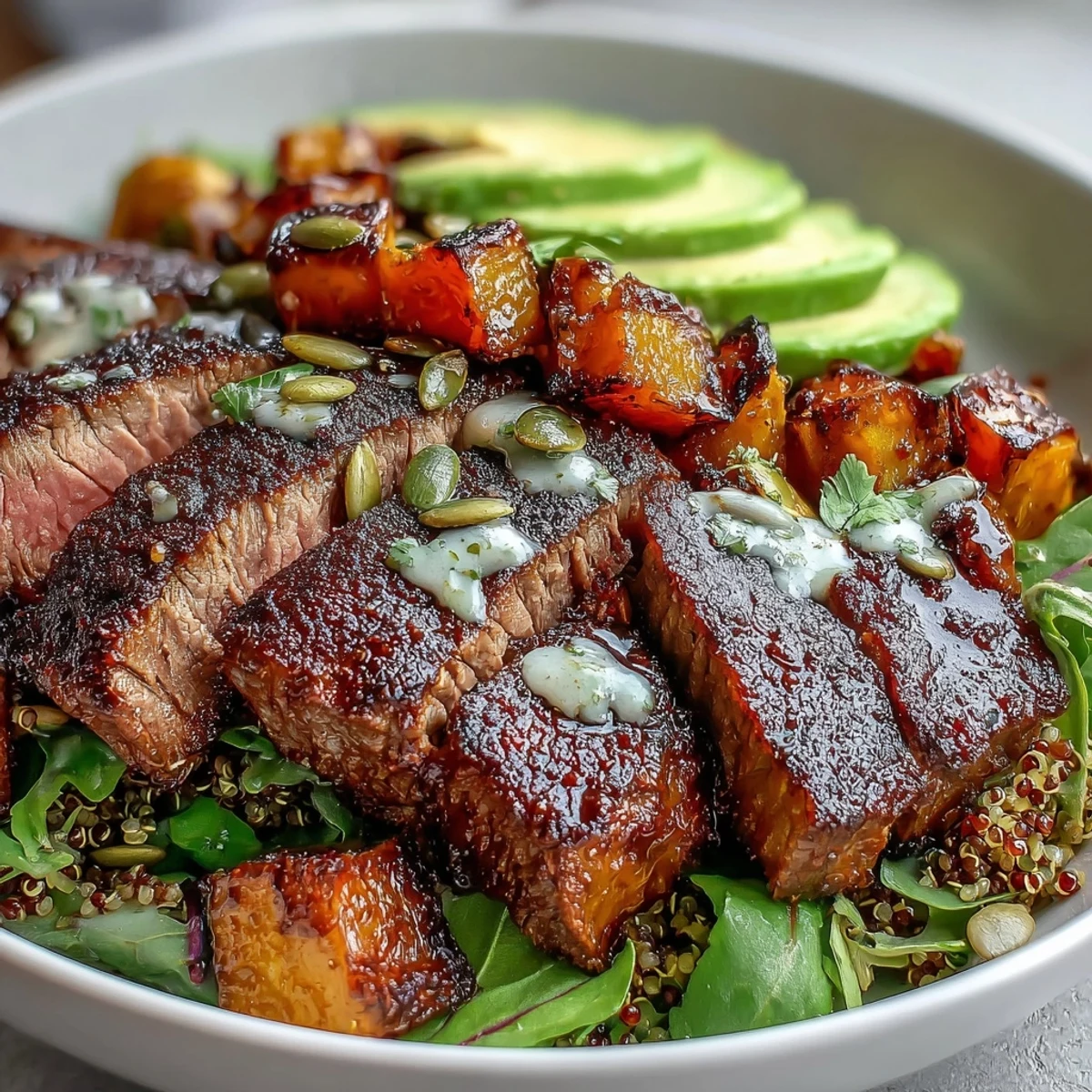 Sliced smoky steak and caramelized butternut squash steak bowls are garnished with red onion, pepitas, and avocado, resting on a bed of greens and warm quinoa.