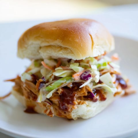 Golden BBQ chicken sliders on toasted buns topped with creamy crunchy slaw, served on a wooden board.