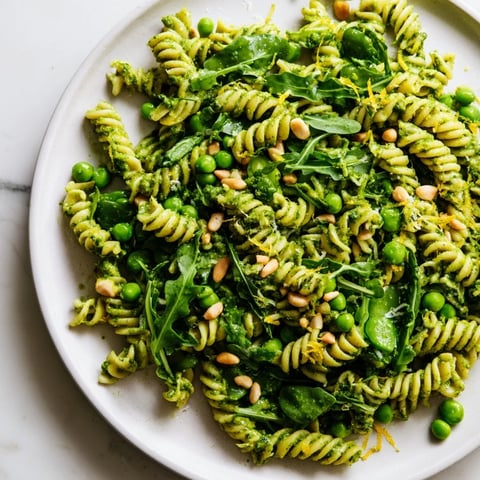 A close-up of vibrant Spring Green Pesto Pasta Salad with fusilli, peas, and arugula tossed in bright green basil pesto and topped with toasted pine nuts.
